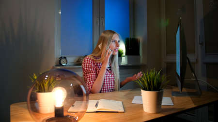 Sitting in dark room lit by small lamp at wooden desk with notebook, young blond woman in checkered shirt talking on mobile phone and looking stunned at computer monitor.の写真素材