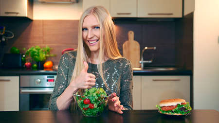 Young lady preferring salad to hamburger. Attractive young woman choosing to eat healthy salad for lunch while sitting at table in stylish kitchen and doing thumbs up gesture and looking at camera.の写真素材