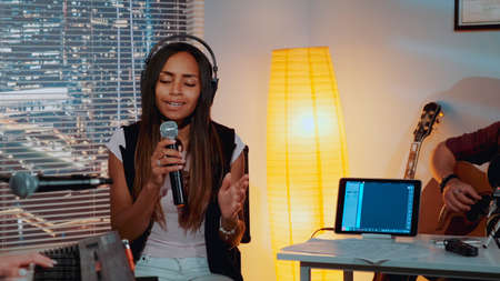 Attractive black woman with close eyes emotionally singing song into microphone and gesturing while her band playing the instruments in home studioの写真素材