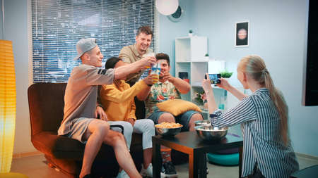 Woman making photo of friends sitting on sofa with cocktails by smartphone. The group of people cheers, having fun and posing on camera.の写真素材