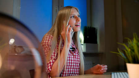 Beautiful smiling blond female in casual checkered shirt sitting at wooden desk with computer at night and looking excitedly at monitor talking on mobile phoneの写真素材