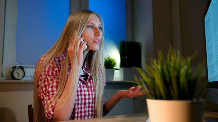 Attractive blond female in casual shirt, sitting at wooden desk and looking with open mouth at computer monitor talking on mobile phone in dark room at nightの写真素材