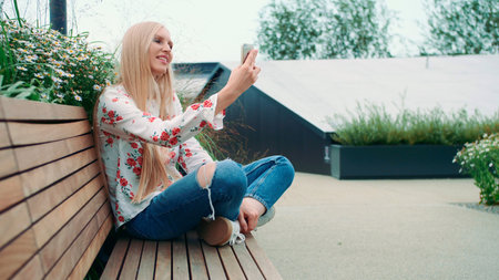 Woman making video call on green roof. Lovely young female using smartphone to make video call while sitting on bench on living roof of huge mall in USA.の写真素材