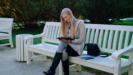 Young Beautiful Woman Sits On The Bench In Autumn Park And Works On The Tablet.の写真素材