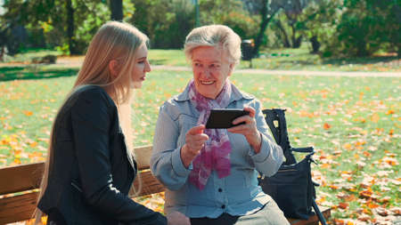 Elder grandmother showing her granddaughter photos on smartphone. They sitting on bench and talking in park.の写真素材