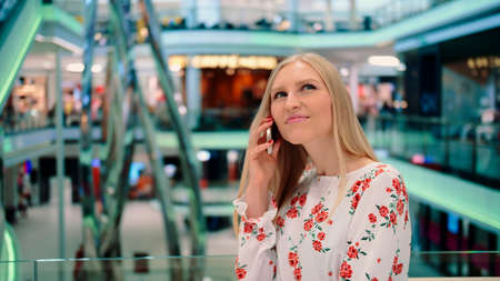 Young woman speaking on phone in mall. Beautiful young woman talking on smartphone and admiring hi-tech interior while standing inside huge mall in Europe.の写真素材
