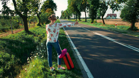 Young woman hitchhiking on countryside road. Back view of young female with red suitcase standing on roadside and hitchhiking while travelling through countryside.の写真素材