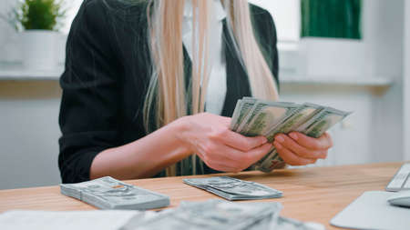 Crop view of female in elegant suit sitting at wooden desk and counting large bundle of dollar banknotes in handsの写真素材