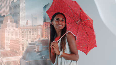 Close-up of smiling black model posing with red umbrella on bar high chair for fashion magazine photo shoot. There are skyscrapers in the background.の写真素材