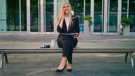 Modern businesswoman in suit sitting on bench with notepad in hands and having phone call looking awayの写真素材