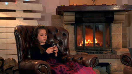 Small girl in nice dress chatting with friends by smartphone in the evening at home. In the background there is a fireplace.の写真素材