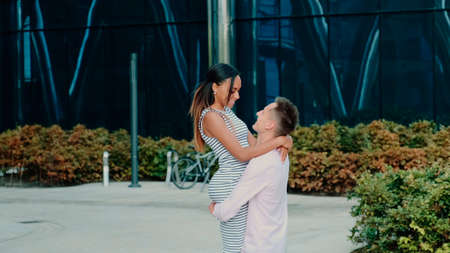 Multiracial family concept: husband spinning around his wife while walking down the street near big office centre one summer dayの写真素材