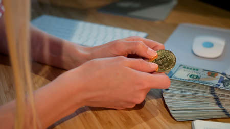 Crop view of hands of woman holding shiny metal bitcoin sitting at wooden desk with wad of cash nearby at nightの写真素材