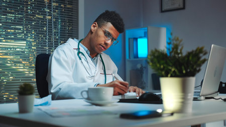 Mixed-race doctor making notes and talking by video call on computer. He working home because of  pandemic.の写真素材
