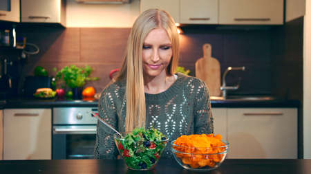 Young lady preferring salad to crisp. Beautiful young woman choosing to eat healthy crisp for supper while sitting at table in stylish kitchen and she eats these salad.の写真素材