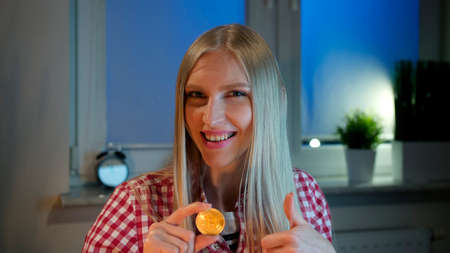 Cheerful young blond woman in casual clothes sitting at dark window at night holding bitcoin and doing thumbs up gesture smiling and looking at cameraの写真素材