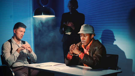 Medium shot of Black prisoner in handcuffs having interview with caucasian detective in interrogation room. Female assistant making notes of the interrogation.の写真素材