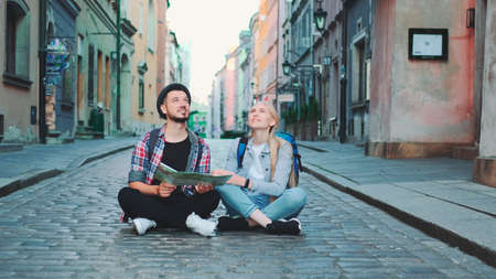 Young tourists couple using map, sitting on pavement and admiring historical surroundings. They are excited and smiled.の写真素材