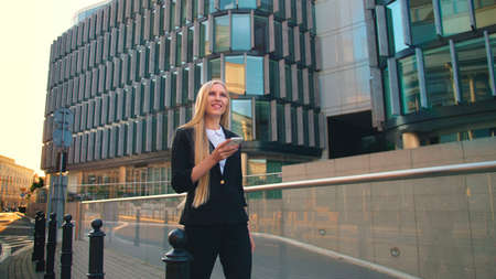 Businesswoman using phone and walking on paved street of contemporary city with new buildings of offices in sunlightの写真素材
