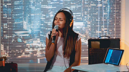 Stylish mixed-race girl singing into microphone at the rehearsal in home studio with skyscrapers in the background.の写真素材