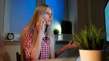 Attractive blond female in casual shirt, sitting at wooden desk and looking with open mouth at computer monitor talking on mobile phone in dark room at night.の写真素材