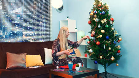 Pretty girl hangs toys on Christmas tree with skyscrapers on the background and sits on sofa with pillows at home. She takes the toys from the box.の写真素材