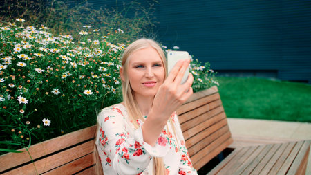 Woman making video call on green roof. Lovely young female using smartphone to make video call while sitting on bench on living roof of huge mall in USA.の写真素材