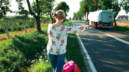 Traveler woman hitchhiking on a sunny road and walking. Young happy backpacker woman looking for a ride to start a journey on a sunlit country road.の写真素材