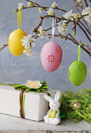 Easter colorful eggs hanging at a blooming branch and gift with easter rabbit on a white wooden tableの写真素材