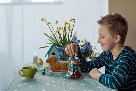 A boy is siting at the table with Easter cakes, eggs and easter decor at homeの写真素材