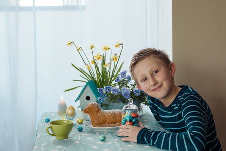 A boy is siting at the table with Easter cakes, eggs and easter decor at homeの写真素材