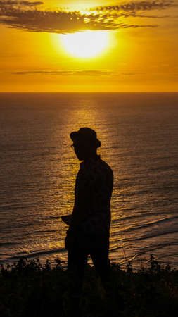 Silhouette of man wearing hat against the background of the sun setting in the ocean.の写真素材