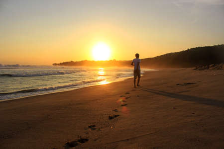 A man walks toward the setting sun, leaving footprints in the sand. Far look from behind.の写真素材