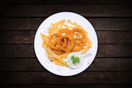 Onion rings and fries with tartar sauce served on a white plate on a dark table, top view.