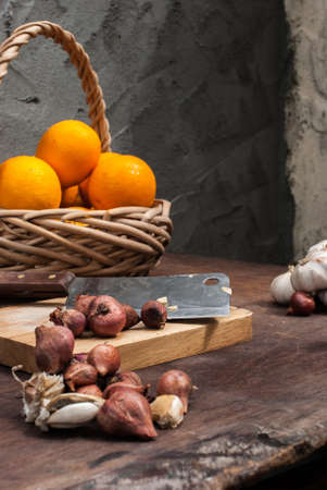 Shallots , Onion , Garlic , Chopping board , Knife , Orange with basket on the table