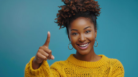 Radiant young African woman in yellow sweater pointing upwards, joyful expressionの素材