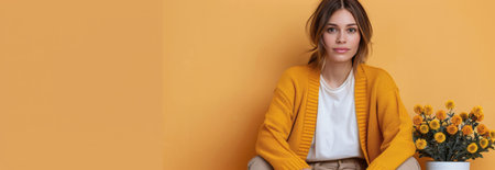 A woman with long brown hair is wearing a yellow cardigan and a white t-shirt. She is sitting in front of a yellow wall. There is a vase of yellow flowers to the right of her.の素材