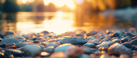 Smooth pebbles are scattered along the shore as the sun sets, casting a warm golden glow over the water, creating a tranquil atmosphere filled with soft light and gentle waves.の素材