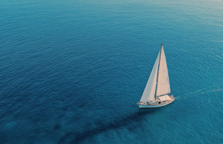 A sailboat navigates the sparkling turquoise sea, leaving a gentle wake as it moves gracefully across the water on a sunny day, highlighting the serenity of the moment.の素材