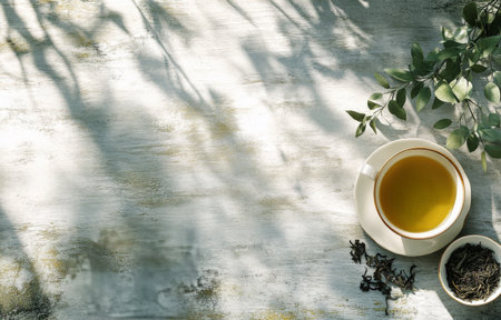 A cup of green tea rests on a wooden table surrounded by loose tea leaves and fresh leaves. Gentle light casts soft shadows, creating a peaceful ambiance for enjoying this calming beverage.の素材
