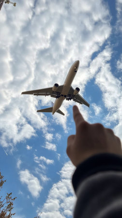 Under a bright blue sky, an individual stretches their hand towards an airplane soaring overhead, surrounded by fluffy white clouds. It captures the spirit of travel and adventure.の素材
