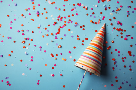 A party hat lies on a vibrant blue surface, surrounded by a mix of colorful confetti. The setting suggests a joyful celebration, perfect for birthdays or special events.の素材