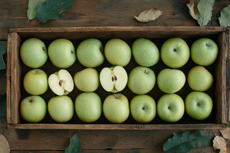 A rustic wooden crate holds a collection of vibrant green apples, with one apple cut in half. The setting has soft, muted lighting that enhances the natural beauty of the fruit.の素材