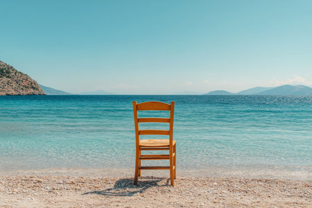 A wooden chair is positioned on a sandy beach, facing the peaceful ocean. The clear skies enhance the tranquil atmosphere, inviting relaxation and reflection by the water.の素材