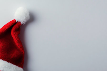 A bright red Christmas hat with a fluffy white pom pom is positioned on a smooth gray surface, ready to bring holiday cheer and warmth to celebrations and gatherings.の素材