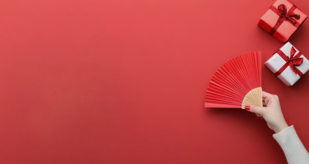 A hand holds a vibrant red fan next to beautifully wrapped gift boxes adorned with ribbons. Decorative branches with small red berries add a festive touch to the soft pink background.の素材