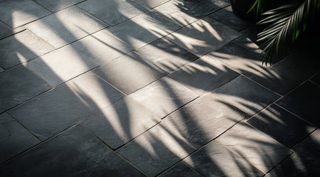Soft shadows of palm leaves create intricate patterns on dark stone tiles during a sunny afternoon. The play of light adds depth and warmth to the outdoor setting.の素材