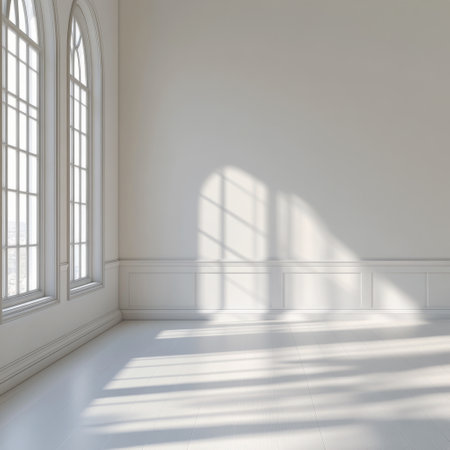 Natural light streams through expansive windows, casting soft shadows on the white walls of a spacious, minimalist room. A hint of greenery is visible outside, enhancing the serene atmosphere.の素材
