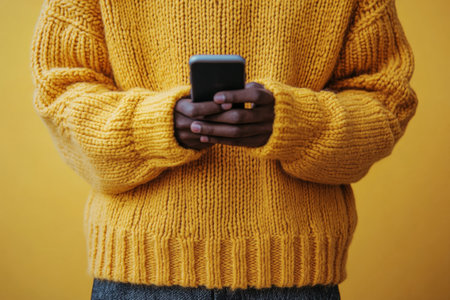 A man dressed in a cozy yellow sweater is attentively looking at his smartphone. Surrounded by vibrant greenery, he appears focused on his device in the bright daylight.の素材