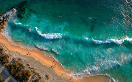 Surfers ride the waves while beachgoers stroll along the shore at a picturesque coastline during sunset. The vibrant water reflects the warm glow of the fading sun.の素材